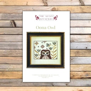 May include: A counted cross stitch pattern for an owl with a brown, white, and red design. The pattern is titled "Oona Owl" and is by The Artsy Housewife. The pattern is framed in a black frame with a gold trim.