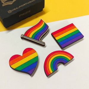 May include: A black box with the word "Pride" in gold lettering, along with four rainbow-colored wooden pins in the shape of a flag, a square, a heart, and a rainbow. The box has the Instagram handle @HBA.PORTUGAL and the word "PRIDE".