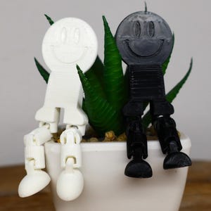 以下が含まれることがあります： 白い笑顔の顔と黒い笑顔の顔の2つの関節式フィギュアが、緑の植物が入った白い鉢に座っています。フィギュアは、笑顔の顔、長方形の体、関節のある脚を持つ丸い頭をしています。