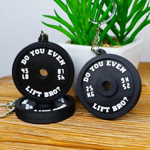 Peut inclure: Porte-clés noirs en forme de disque de poids avec le texte "DO YOU EVEN LIFT BRO ?" et des mesures de poids en kilogrammes (25 kg). Un porte-clés affiche 45 lbs et 81 lbs. Sur une surface en bois.