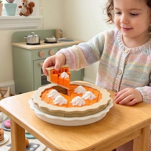 May include: A wooden toy pie set featuring a tan crust and orange filling, topped with white dollops of whipped cream. The set includes a white serving dish and individual pie slices that can be lifted, designed for interactive play and learning.