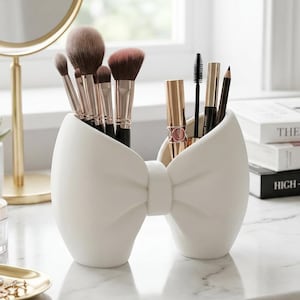 May include: A white bow-shaped makeup brush holder filled with brushes, mascara, and eyeliner. The holder is on a marble surface, near a gold-framed mirror and a small succulent plant. The overall aesthetic is clean and elegant.
