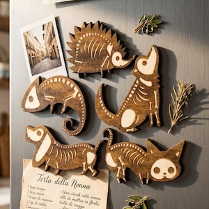 Puede incluir: Imanes de madera con forma de esqueletos de animales, como un erizo, un perro, un gato y un camaleón, se exhiben en un refrigerador. También se adjunta una tarjeta de receta italiana. Un dibujo infantil es visible.
