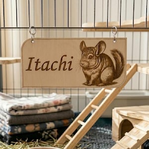 Può includere: Un cartello di legno con il nome "Itachi" e un'illustrazione dettagliata di un cincillà. Il cartello è appeso all'interno di una gabbia per cincillà, che comprende piattaforme in legno, una scala e una casetta. La gabbia contiene anche lettiera, cibo e una pila di coperte.