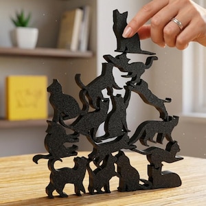 Peut inclure: Jeu d'équilibre en forme de chat noir en cours d'assemblage. Le jeu se compose de plusieurs silhouettes de chats noirs empilées les unes sur les autres. Le jeu est sur une table en bois. Une main place une silhouette de chat sur le dessus de la pile.