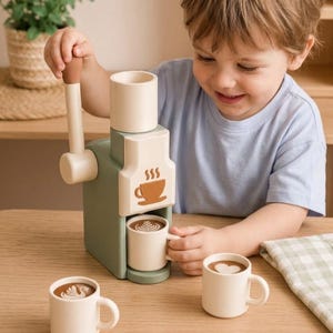 May include: A sage green and cream toy coffee machine with a manual lever, featuring a brown coffee cup icon on the front. The set includes three small cream-colored mugs, each displaying a different latte art design: a leaf, a swan, and a heart.