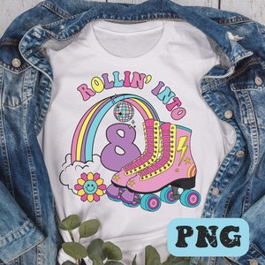 Peut inclure: Un t-shirt blanc avec une impression graphique portant le texte ROLLIN' INTO, un grand chiffre 8 violet, un arc-en-ciel, une boule à facettes, un éclair jaune et des patins à roulettes roses à roues bleues, sous une veste en jean bleu.