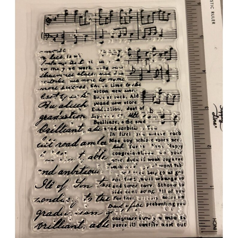 May include: Clear, rectangular stamp with musical notes and handwritten script. The script includes words like "graduation" and "brilliant." A ruler with inch measurements is visible on the right side of the stamp.