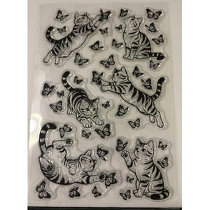 Puede incluir: Una hoja transparente de sellos de fotopol&iacute;mero con ilustraciones de l&iacute;neas negras de cuatro gatos atigrados en varias poses, incluyendo saltando, sentados y jugando, rodeados de numerosos dise&ntilde;os de mariposas sobre la superficie.