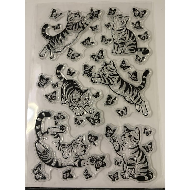 Puede incluir: Una hoja transparente de sellos de fotopol&iacute;mero con ilustraciones de l&iacute;neas negras de cuatro gatos atigrados en varias poses, incluyendo saltando, sentados y jugando, rodeados de numerosos dise&ntilde;os de mariposas sobre la superficie.