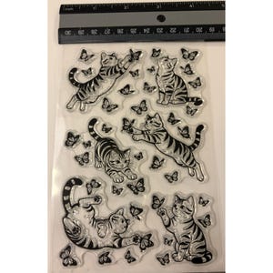 Puede incluir: Una hoja transparente de sellos de silicona con cinco gatos atigrados de contorno negro en varias posturas rodeados de mariposas. Una regla negra est&aacute; colocada en la parte superior, mostrando medidas en cent&iacute;metros, con unos 10 cent&iacute;metros de ancho.
