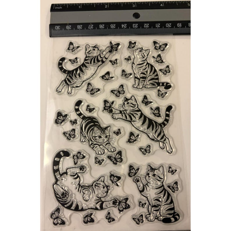 Puede incluir: Una hoja transparente de sellos de silicona con cinco gatos atigrados de contorno negro en varias posturas rodeados de mariposas. Una regla negra est&aacute; colocada en la parte superior, mostrando medidas en cent&iacute;metros, con unos 10 cent&iacute;metros de ancho.
