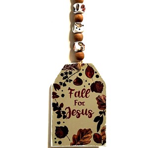 May include: A beige wooden tag with a fall-themed design and the text "Fall For Jesus" in red lettering. The tag is attached to a brown and white beaded string.