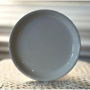 May include: A round, off-white ceramic plate with a slightly raised edge. The plate has a smooth, glossy finish and is set against a neutral background. The plate is likely for serving food.