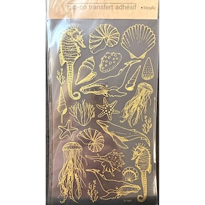 May include: Gold metallic rub-on transfer sheet with a variety of sea life designs, including seahorses, whales, jellyfish, starfish, and seashells. The sheet is labeled "rub-on transfert adhésif Metallic".