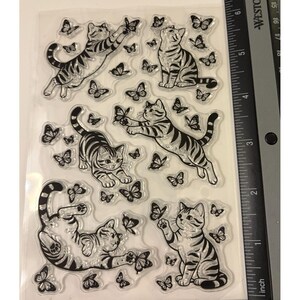 Puede incluir: Una hoja de sellos de silicona transparente con cinco ilustraciones distintas de gatos atigrados interactuando con varias mariposas. Una regla negra Westcott est&aacute; colocada en el borde derecho, mostrando medidas en cent&iacute;metros hasta 15 cm.