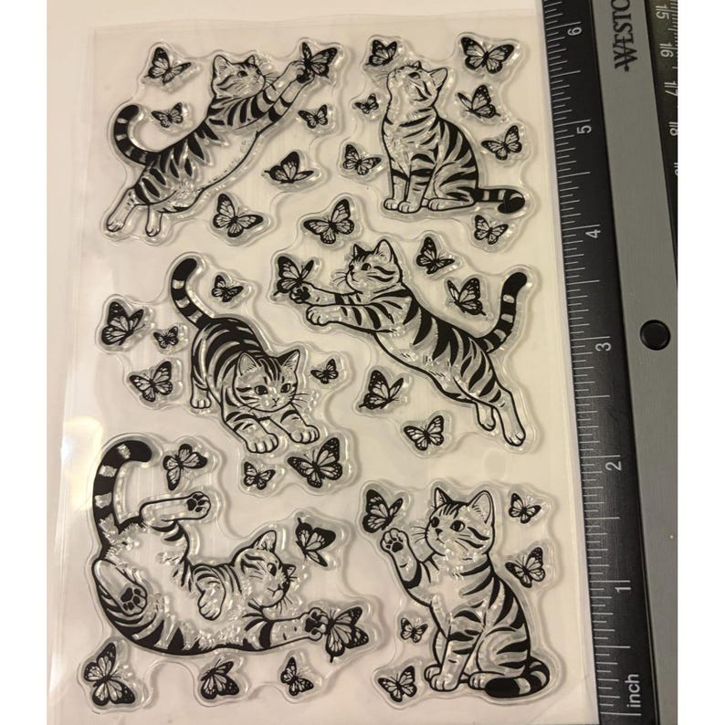 Puede incluir: Una hoja de sellos de silicona transparente con cinco ilustraciones distintas de gatos atigrados interactuando con varias mariposas. Una regla negra Westcott est&aacute; colocada en el borde derecho, mostrando medidas en cent&iacute;metros hasta 15 cm.