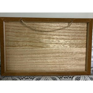 May include: A rectangular wooden sign with a light brown wood grain pattern. The sign has a dark brown frame and a natural fiber rope for hanging. The wood grain runs horizontally across the sign.