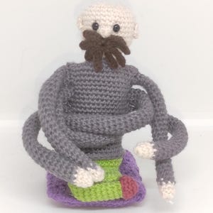 Puede incluir: Muñeca de ganchillo hecha a mano con cabeza beige claro, bigote marrón y cuerpo gris con múltiples brazos. La muñeca tiene una base morada con detalles verdes y rojos. La muñeca está sobre un fondo blanco.