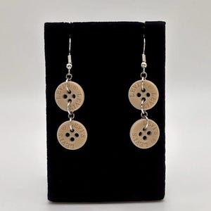 May include: Beige button earrings with the word "LEVI'S" printed around the edge of each button. The earrings are made of two buttons connected by a silver chain and hang from silver hooks. The earrings are displayed on a black surface.