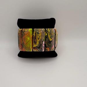 May include: A colorful bracelet with a marbled design. The bracelet features rectangular, chunky links in shades of yellow, pink, brown, and black. The bracelet is displayed on a black velvet stand against a white background.