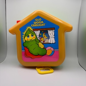 May include: A vintage Glo Worm Criblight toy in the shape of a house. The orange plastic house features a blue window with a cartoon worm character, a moon, and stars. The toy has a yellow handle and a red button.