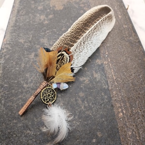 Puede incluir: Una pluma marrón y blanca con un alambre de cobre envuelto alrededor. La pluma tiene un pequeño amuleto atrapasueños con cristales y una pluma blanca adjunta.