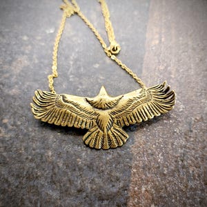 May include: A gold-toned metal necklace featuring a pendant shaped like an eagle with its wings spread wide. The pendant displays detailed etched feather patterns across the wings and tail. The piece is attached to a thin, matching gold-toned chain.