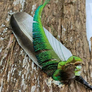 May include: A decorative feather quill featuring a large, grey-brown base feather layered with a vibrant, iridescent green peacock feather. The quill base is wrapped in dark thread and accented with small, pale green beads near the quill shaft.
