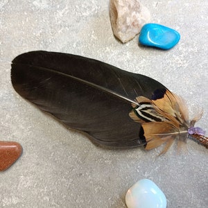 May include: A large, dark feather with brown and tan accents, adorned with a wire-wrapped amethyst crystal. The feather is surrounded by various polished stones in shades of white, orange, and blue, set against a gray background.