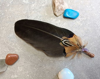 Puede incluir: Una pluma grande de color negro decorada con plumas más pequeñas de rayas marrones y blancas en la base. El cañón está envuelto en alambre de cobre y rematado con un pequeño cristal morado. La pluma está rodeada por cuatro piedras pulidas.