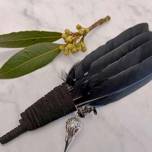 Puede incluir: Pluma de plumas negras hecha a mano con un dije de plata decorativo con diseño de calavera. La pluma va acompañada de hojas de laurel verdes y una ramita de bayas de color amarillo verdoso, sobre un fondo de mármol blanco.