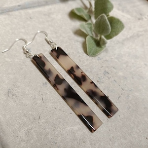 Puede incluir: Un par de pendientes largos de plata con colgantes rectangulares de color marrón y blanco con estampado de caparazón de tortuga.