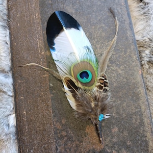 Könnte beinhalten: Eine Pfauenfeder mit einem blauen und grünen Auge, akzentuiert mit braunen und weißen Federn und einer türkisfarbenen Perle an der Basis.