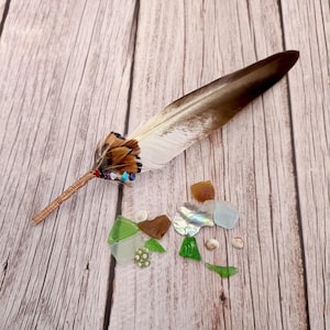 Puede incluir: Una pluma decorativa con tonos marrones y blancos, adornada con cuentas de colores y alambre de cobre. Acompañando a la pluma hay trozos de vidrio marino en varios colores, incluyendo verde, marrón y azul, junto con pequeñas conchas.