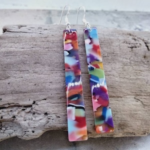 May include: A pair of long, colourful dangle earrings. The earrings are made of a rectangular piece of colourful, translucent plastic with a silver hook.