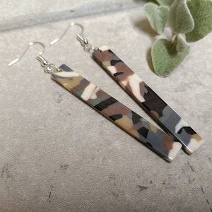 May include: A pair of dangle earrings with silver hooks and long rectangular dangles featuring a camouflage pattern in shades of grey, brown, and white.