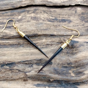 May include: A pair of black and gold earrings with a pointed design. The earrings are made of wood and metal.