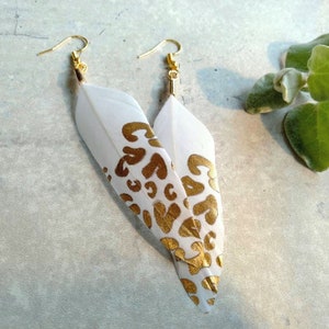 May include: A pair of white feather earrings with gold leopard print designs. The earrings have gold hooks.