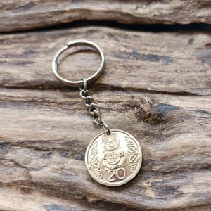 Puede incluir: Un llavero con una moneda de color bronce, un llavero y una cadena plateados. La moneda presenta un relieve detallado de una figura y el número 20. El llavero se muestra sobre un fondo de madera.