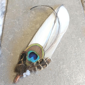 May include: A decorative feather accessory featuring a large white primary feather layered with a smaller peacock eye feather. The base is wrapped in copper-coloured wire and accented with a small, translucent white crystal bead.