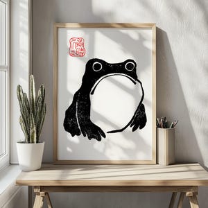 May include: Black and white print of a frog on a white background with a red stamp in the top left corner. The stamp says "蛙" in Japanese characters.