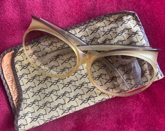 Puede incluir: Gafas ovaladas de color beige translúcido con silueta de ojo de gato, apoyadas sobre un estuche de tela con estampado beige y negro. El estuche presenta un patrón repetido del logotipo YSL y el texto Saint Laurent en el borde lateral.