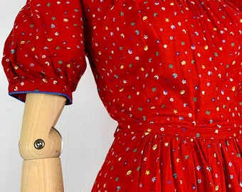 May include: A red dress with a white floral print and blue trim. The dress has a Peter Pan collar and short puffed sleeves. The dress is cinched at the waist with a belt.