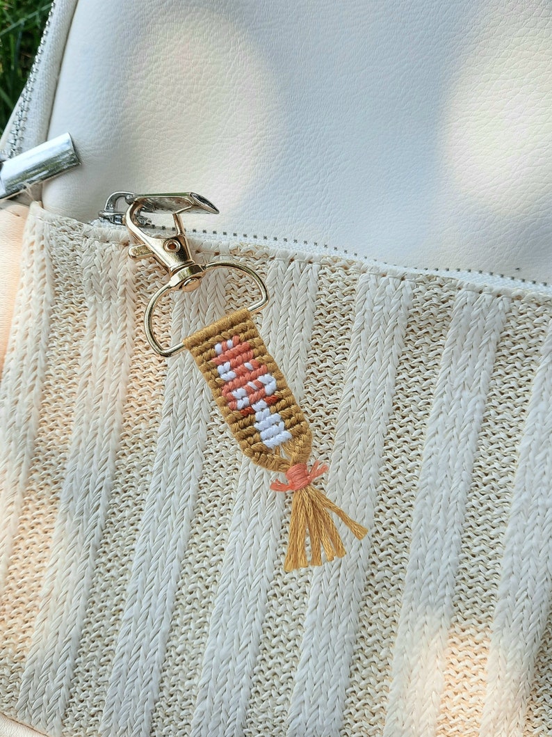 K&ouml;nnte beinhalten: Ein handgefertigter Schl&uuml;sselanh&auml;nger mit einem Pilzdesign. Der Anh&auml;nger besteht aus gewebten braunen, wei&szlig;en und pfirsichfarbenen F&auml;den, mit einem goldfarbenen Verschluss und einer Quaste aus beigefarbenen F&auml;den.
