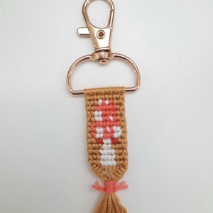 K&ouml;nnte beinhalten: Handgefertigter Schl&uuml;sselanh&auml;nger mit einem gewebten Pilzdesign in Pfirsich, Wei&szlig; und Braun. Der Schl&uuml;sselanh&auml;nger hat einen goldfarbenen Verschluss und eine Quaste aus braunem Garn mit einem pfirsichfarbenen Akzent.