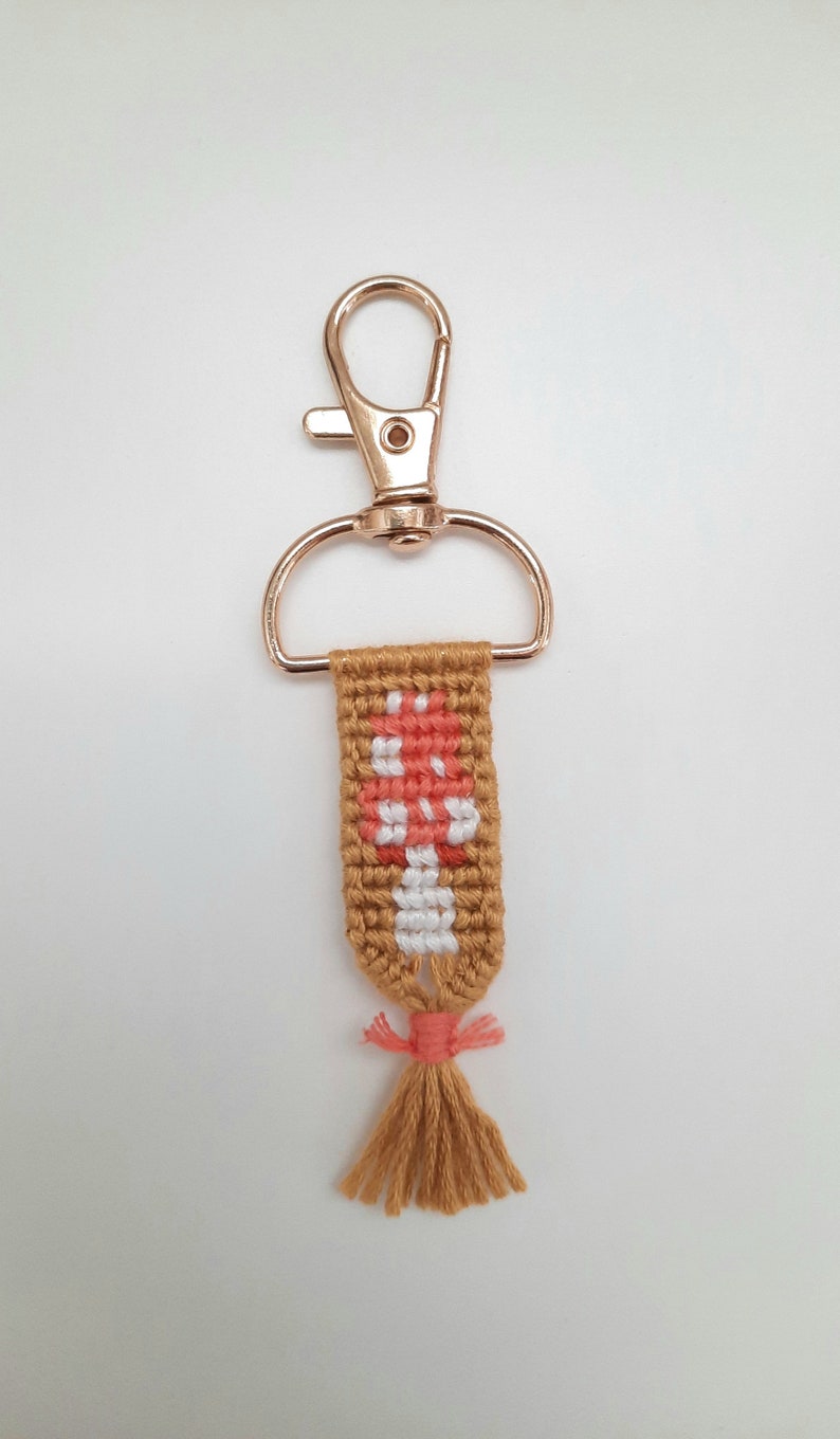 K&ouml;nnte beinhalten: Handgefertigter Schl&uuml;sselanh&auml;nger mit einem gewebten Pilzdesign in Pfirsich, Wei&szlig; und Braun. Der Schl&uuml;sselanh&auml;nger hat einen goldfarbenen Verschluss und eine Quaste aus braunem Garn mit einem pfirsichfarbenen Akzent.
