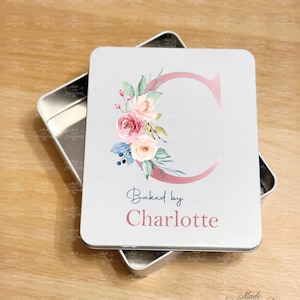 May include: A white metal tin with a pink floral design and the text "Baked by Charlotte". The tin is closed and the lid is sitting on top of the tin. The lid has a pink floral design with a pink letter "C" in the center. The tin is made to be personalised.