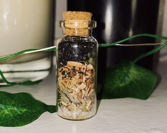 May include: A small glass bottle with a cork stopper filled with a layered mixture of black, brown, and green herbs and crystals.