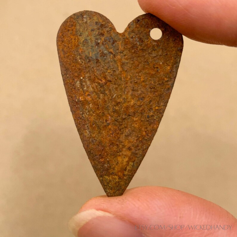 Puede incluir: Un colgante met&aacute;lico en forma de coraz&oacute;n con una superficie oxidada y texturizada. El colgante tiene un peque&ntilde;o agujero en la parte superior para colgarlo. El color es principalmente marr&oacute;n con toques de azul y verde. La URL de la tienda Etsy es visible en la parte inferior.
